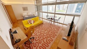 Un salon moderne avec un canapé jaune, un tapis à motifs, une table à manger et une grande fenêtre donnant sur des immeubles.