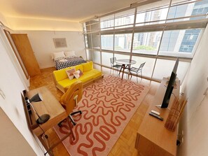 A modern living room with a yellow sofa, a patterned rug, a dining table, and a large window offering a view of buildings. - 0517B (São Paulo)