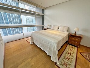 A bedroom with a large window, a bed with a white bedspread, a nightstand with a lamp, and a patterned rug. - 0121C (São Paulo)