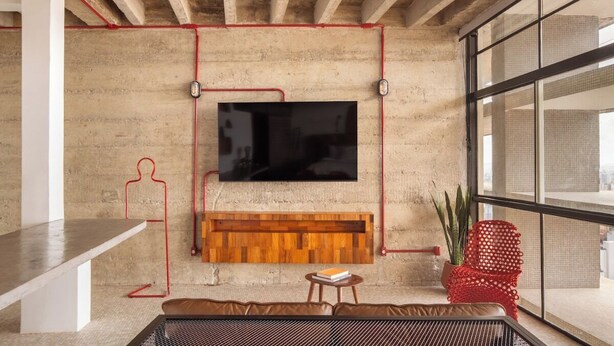 A modern living room with a large flat-screen TV mounted on a concrete wall, a wooden bench below, a red chair, and a small table with a book. - 3220B (São Paulo)