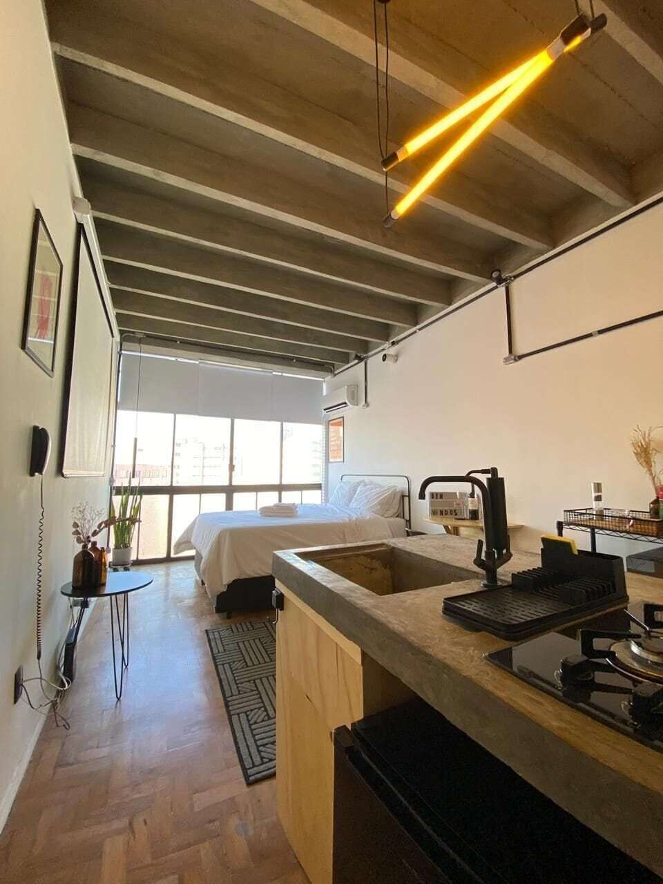 A modern kitchen with a large island, a bed with white linens, and a view of the outdoors through large windows.