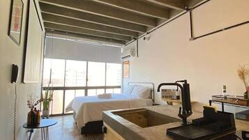A modern kitchen with a large island, a bed with white linens, and a view of the outdoors through large windows.