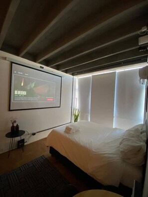 Un dormitorio con cama, televisor montado en la pared y ventana con persianas.