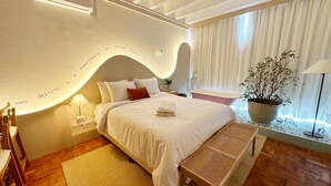 Ein modernes Schlafzimmer mit einer gewölbten Wand, einem Bett mit weißer Bettwäsche, einem Nachttisch mit Lampe, einer Bank und einem Blumentopf am Fenster.