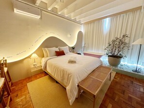 Ein modernes Schlafzimmer mit einer gewölbten Wand, einem Bett mit weißer Bettwäsche, einem Nachttisch mit Lampe, einer Bank und einem Blumentopf am Fenster.