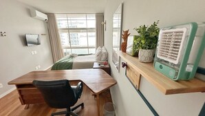 A room with a wooden desk, a black office chair, a small trash bin, a shelf with decorative items, and a wall-mounted air conditioner. - 0055F (São Paulo)