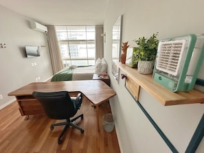 Ein Zimmer mit Schreibtisch aus Holz, schwarzem Bürostuhl, kleiner Mülleimer, Regal mit Dekoration und Wandklimaanlage.