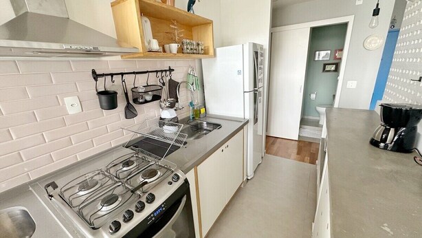 A modern kitchen with a gas stove, oven, and white cabinetry. There is a wooden shelf above the sink with various items. - 0055F (São Paulo)
