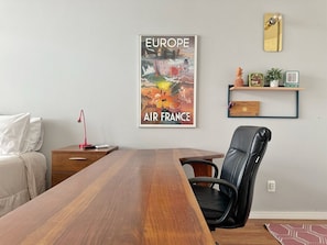 Ein Zimmer mit Holztisch, schwarzem Bürostuhl, Bett mit weißer Bettwäsche, roter Schreibtischlampe, einem Poster von Europa und Air France, einem Regal mit Dekoration und einem Spiegel an der Wand.
