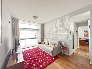 Ein modernes Wohnzimmer mit einem grauen Sofa, einem roten Teppich mit Muster, einer weißen Wand mit Rasteroptik und einem großen Fenster.