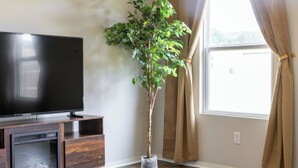Un salon avec un téléviseur à écran plat posé sur un meuble en bois, une plante en pot et une fenêtre avec des rideaux.