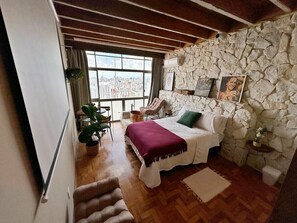 A bedroom with a stone wall, a bed with a white cover, a window with a city view, and a wooden floor. - 2911B (São Paulo)