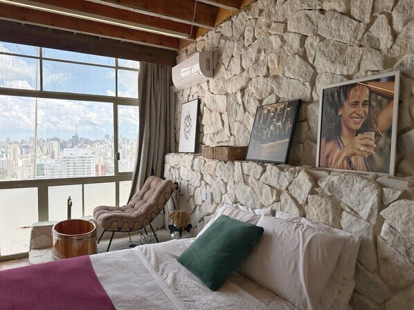 Une pièce avec un mur en pierre, un lit avec un oreiller vert, une chaise, un seau et une vue sur la ville.