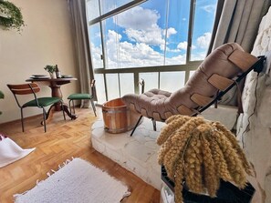 A room with a large window, a cushioned chair, a wooden bucket, and a small table with a plant. - 2911B (São Paulo)