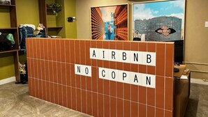 Ein Empfangsbereich mit einer Holzwand, auf der 'AIRBNB NO COPAN' steht, und zwei gerahmten Kunstwerken an der Wand.