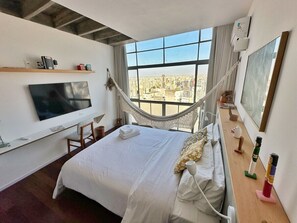 A hotel room with a large bed, a TV, a hammock, and a city view.
