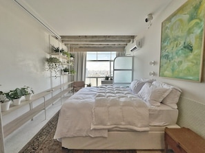 Ein Schlafzimmer mit einem großen Bett, Blick auf die Stadt durch ein Fenster und mehreren Blumentöpfen auf einem Regal.