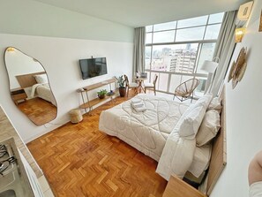 A modern hotel room with a large bed, a TV, a desk with a chair, and a view of the city through the window. - 2407B (São Paulo)