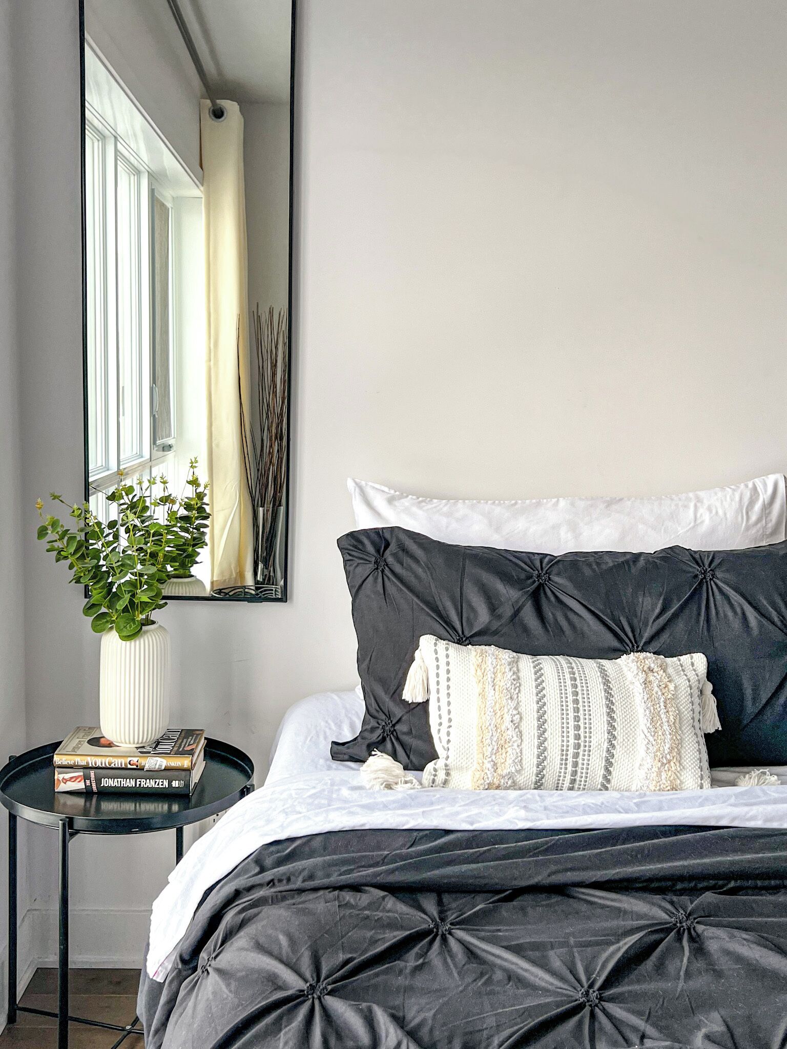 A bedroom with a bed, a bedside table, a mirror, and a potted plant.