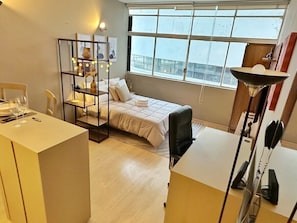 A hotel room with a bed, a desk, a chair, a shelf with decorative items, and a large window.