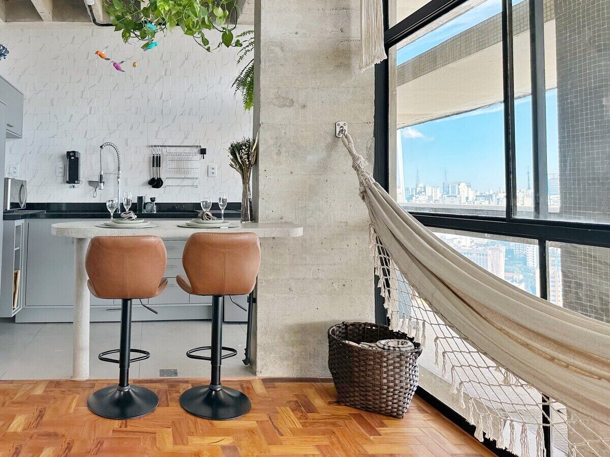 A modern kitchen with a white countertop, two bar stools, a hanging hammock, and a city view through large windows.