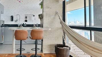 A modern kitchen with a white countertop, two bar stools, a hanging hammock, and a city view through large windows.