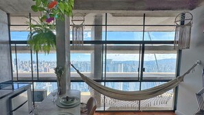 A dining area with a table set for two, a hammock, and a city view through large windows.