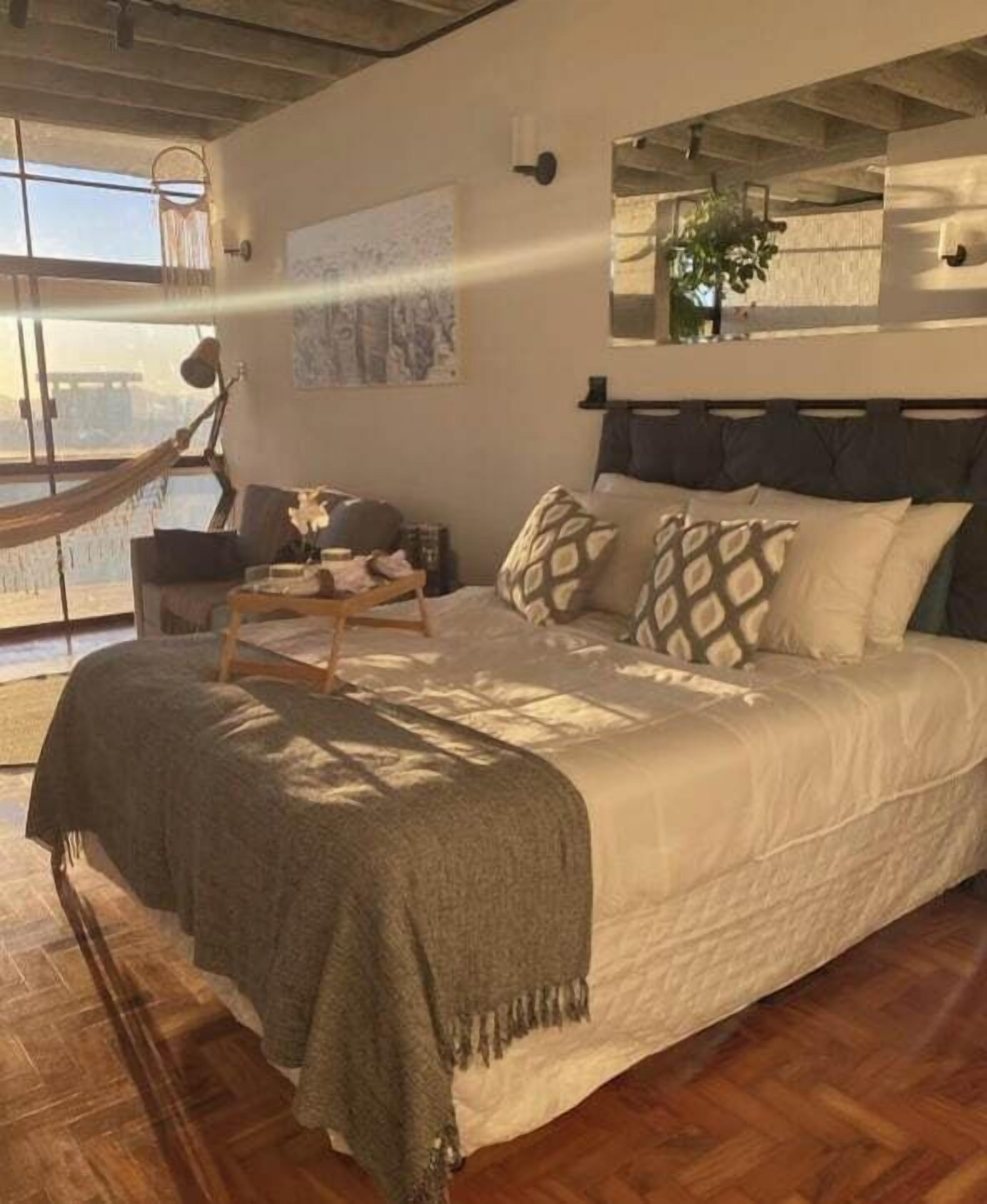 A bedroom with a large bed, a hammock, a view through a window, and a plant on the windowsill.