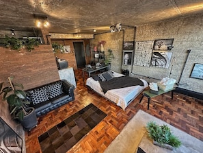 A loft-style bedroom with a herringbone floor pattern, a bed with a white and black bedspread, a black leather sofa, and a small table with a vase. - 3013M (São Paulo)