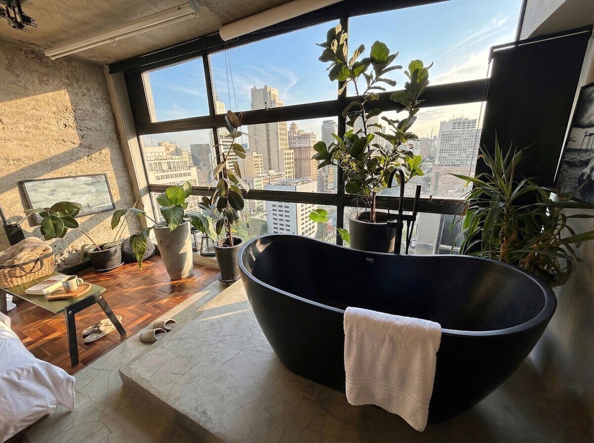Ein modernes Badezimmer mit einer großen schwarzen Badewanne, zahlreichen Topfpflanzen und einem Blick auf die Stadt durch bodentiefe Fenster.