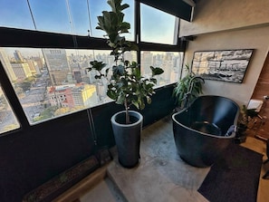 A room with a large window overlooking a cityscape, a potted plant, and a black bathtub. - 3013M (São Paulo)