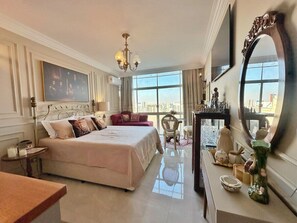A hotel room with a large bed, a sitting area with a sofa, a dining table, and a large window offering a city view. - 2312B (São Paulo)