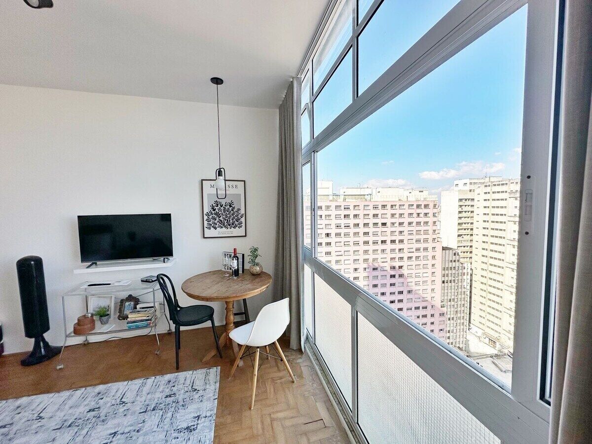 A modern living room with a large window, a flat-screen TV, a round wooden table, and a view of a cityscape.