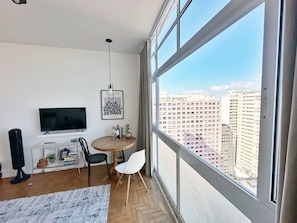A modern living room with a large window, a flat-screen TV, a round wooden table, and a view of a cityscape. - 2011B (São Paulo)