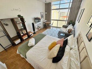 A modern hotel room with a bed, a desk, a chair, a wardrobe, and a large window offering a city view. - 2011B (São Paulo)