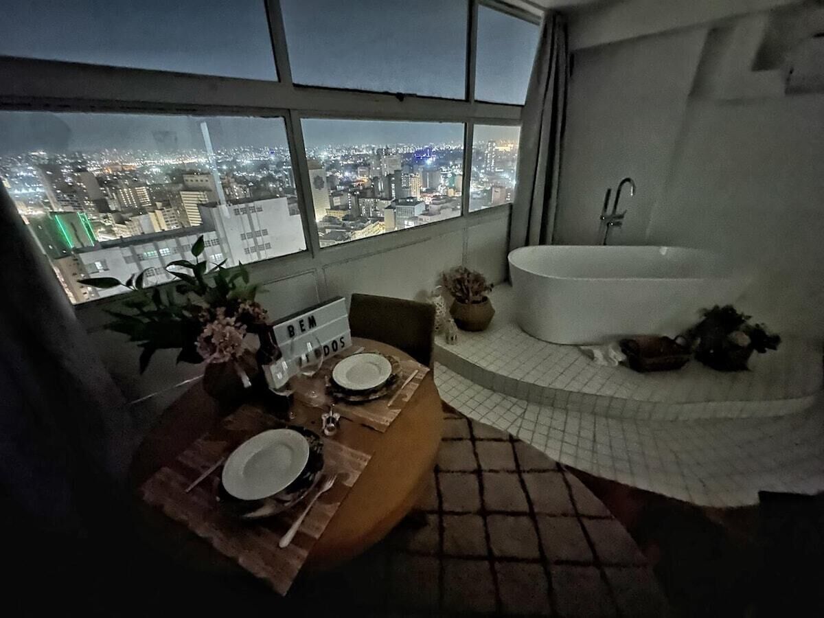 A room with a dining table set for two, a large window with a city view, and a bathtub.
