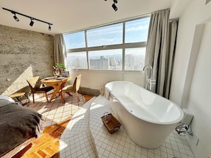 Ein modernes Badezimmer mit freistehender Badewanne, Blick auf die Stadt durch große Fenster und einem Essbereich mit Tageslicht.