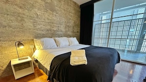 Quarto moderno com uma cama grande, uma mesinha de cabeceira, uma luminária e vista pela janela com grade de metal.