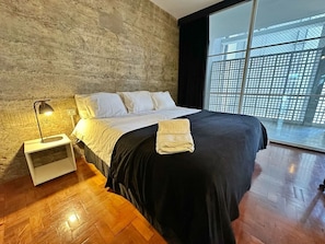 Ein modernes Schlafzimmer mit einem großen Bett, einem Nachttisch, einer Lampe und Blick durch ein Fenster mit Gitterrost.