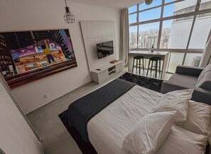 Quarto moderno com cama, televisão, mesa pequena e varanda com vista para a cidade.