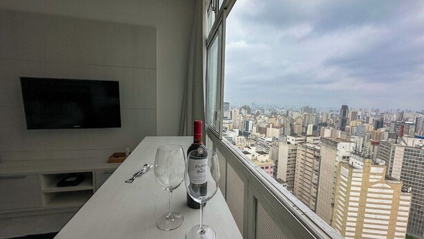 Ein Zimmer mit Tisch am Fenster mit Blick auf eine Stadtlandschaft, einer Weinflasche und einem Fernseher an der Wand.