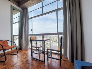 Een kamer met houten vloer, een tafel met stoelen, een bank en een groot raam met uitzicht over de stad.