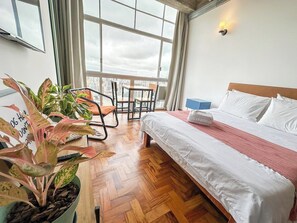 A hotel room with a bed, a desk, a chair, a potted plant, and a view of the city through large windows. - 3205B (São Paulo)