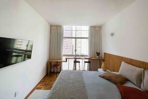 A hotel room with a bed, a TV mounted on the wall, a desk with chairs, and a window with curtains. - 0705B (São Paulo)