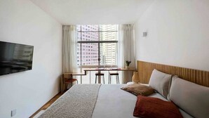 Ett hotellrum med en säng, en tv, ett skrivbord och ett fönster med utsikt över byggnader.