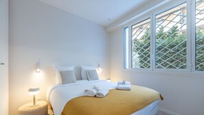 Een net opgemaakt bed met een deken in wit en mosterdgeel, een bedlampje en een raam met uitzicht op groen.