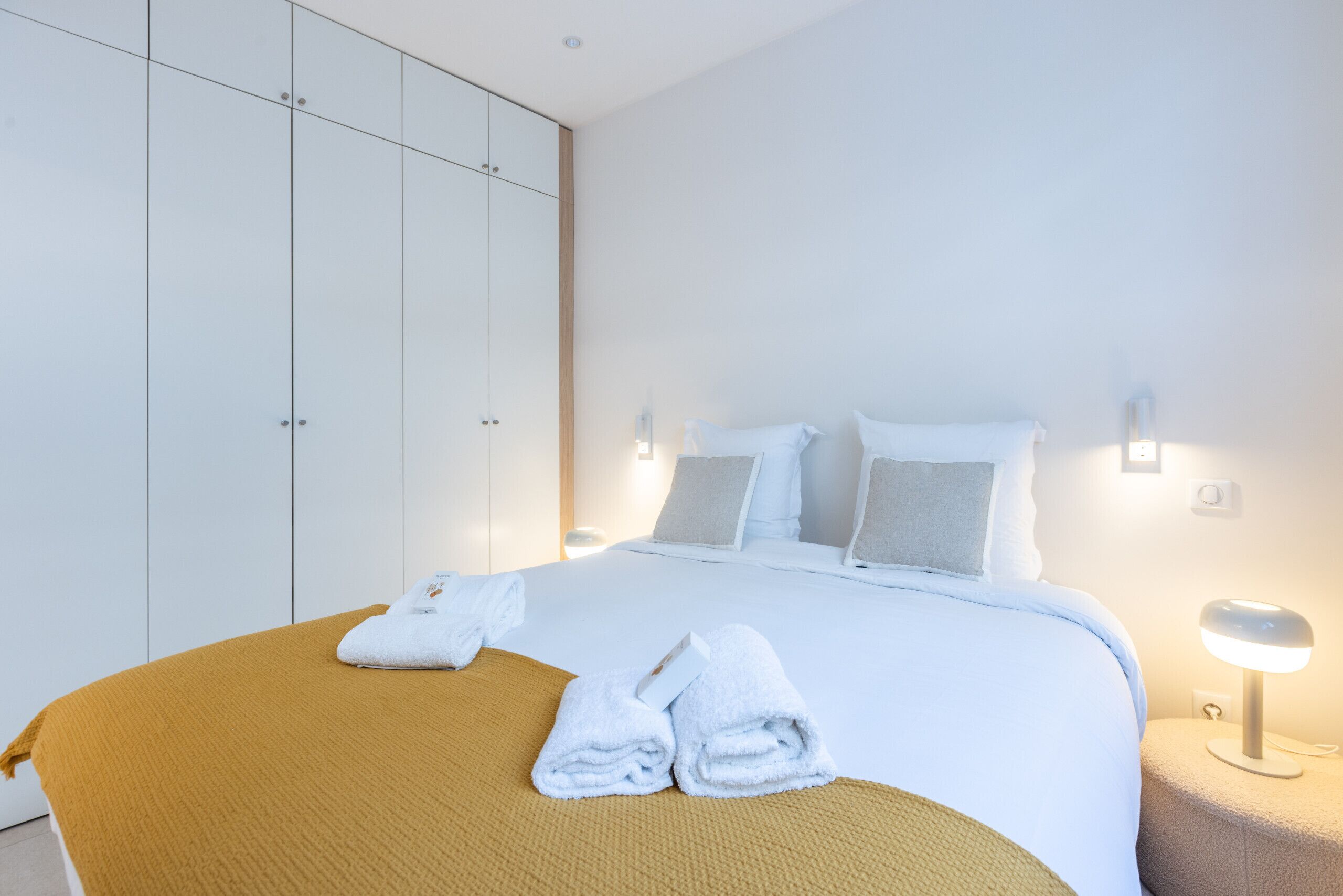A neatly made bed with white linens and a mustard-colored throw blanket. There are two folded towels on the bed. The room has white walls and a large wardrobe.