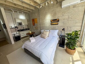 A modern bedroom with a large bed, a small bedside table, a potted plant, and a view of a kitchen area. - 0202B (São Paulo)