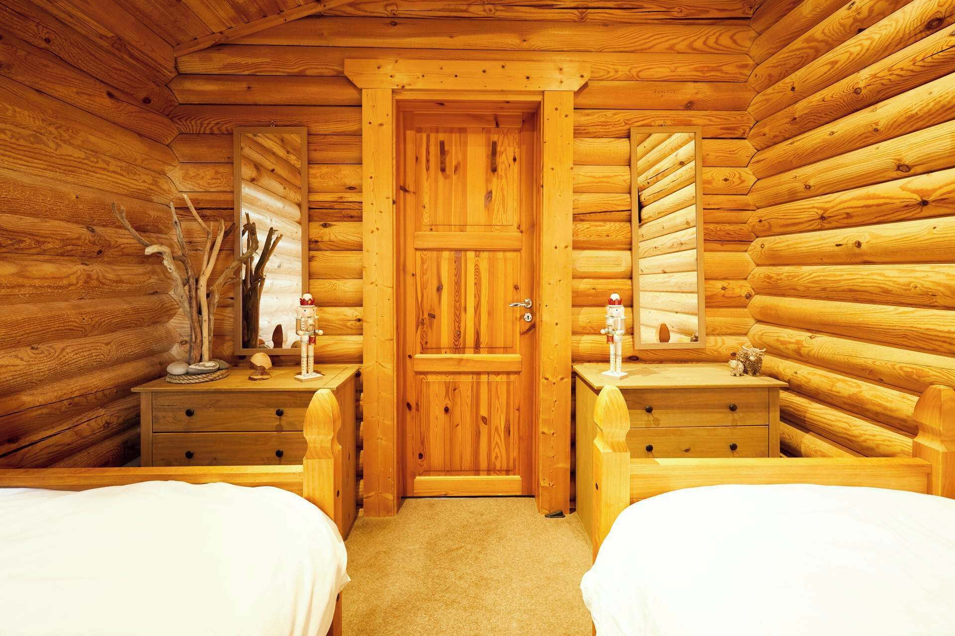 A log cabin bedroom with two beds, wooden walls, a central door, and small bedside tables with decorative items.