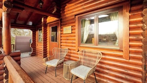 A log cabin with a wooden deck, two wicker chairs, a wooden door, and a window with a view of trees.